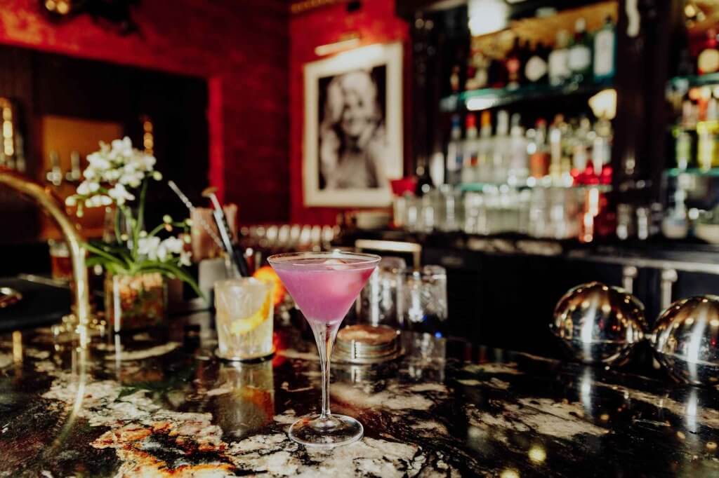 A bar with a pink drink in a martini glass and another drink behind it with an orange garnish.